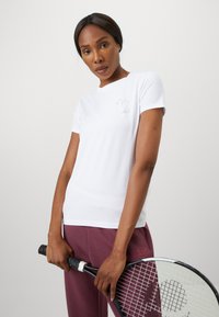 T-shirt bianca a maniche corte realizzata in tessuto liscio, con un piccolo logo argentato sul petto. Il modello tiene in mano una racchetta da tennis nera, indossando pantaloni color bordeaux.