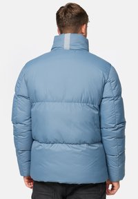 Lys blå jakke med høy krage, quiltet tekstur, elastisk liv og reflekterende detaljer på ryggen.