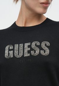 Pull noir avec un col côtelé et un logo "GUESS" bien en vue orné de strass clairs, avec "EST. 1981" imprimé en dessous.