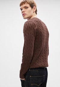 Καφέ πουλόβερ με πλέξη cable-knit και υφή. Έχει στενή εφαρμογή, με στρογγυλή λαιμόκοψη και φοριέται πάνω από σκούρα τζιν.