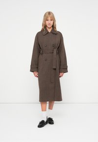 Brun hørgynget trenchcoat med bælte, stort krave og dobbeltbrystet knaplukning; modellen bærer sorte sko og hvide sokker.