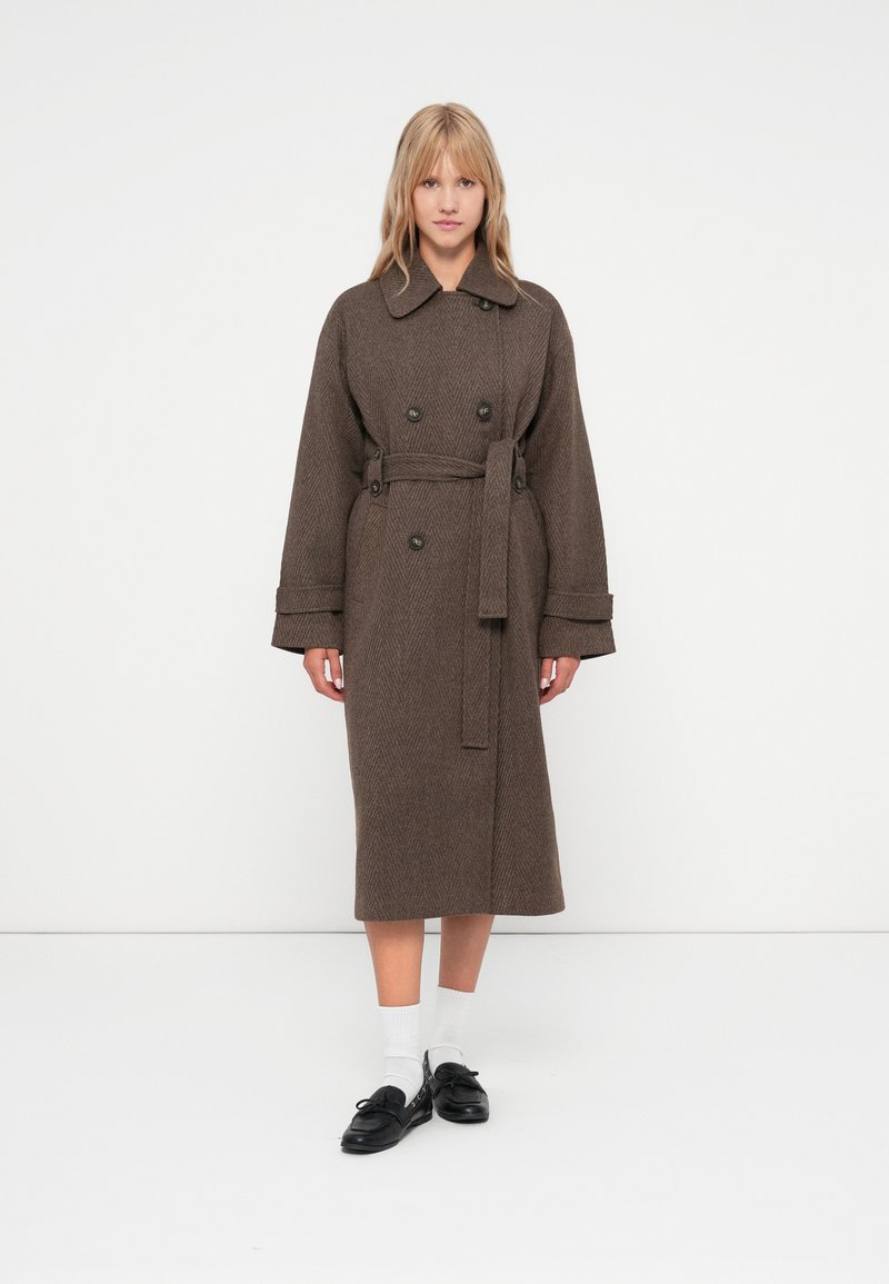 Brun hørgynget trenchcoat med bælte, stort krave og dobbeltbrystet knaplukning; modellen bærer sorte sko og hvide sokker.