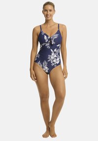 Vrouw in een marineblauwe bloemen ééndelige zwempak, lachend met haar handen op haar heupen, staand op blote voeten tegen een neutrale achtergrond.