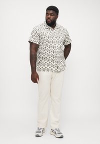 Kurzarmhemd mit Knopfleiste und geometrischem Muster in Beige und Oliv. Kombiniert mit cremefarbenen Hosen und grauen Sportschuhen.