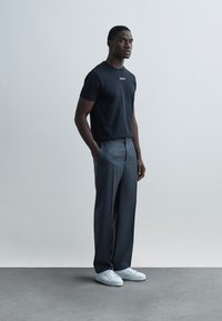 Juoda medvilninė marškinėliai su logotipu, derinami su pilkais languotais kelnėmis. Baltos sportbačiai užbaigia aprangą. Stovinčios pozos su neutraliu fonu.