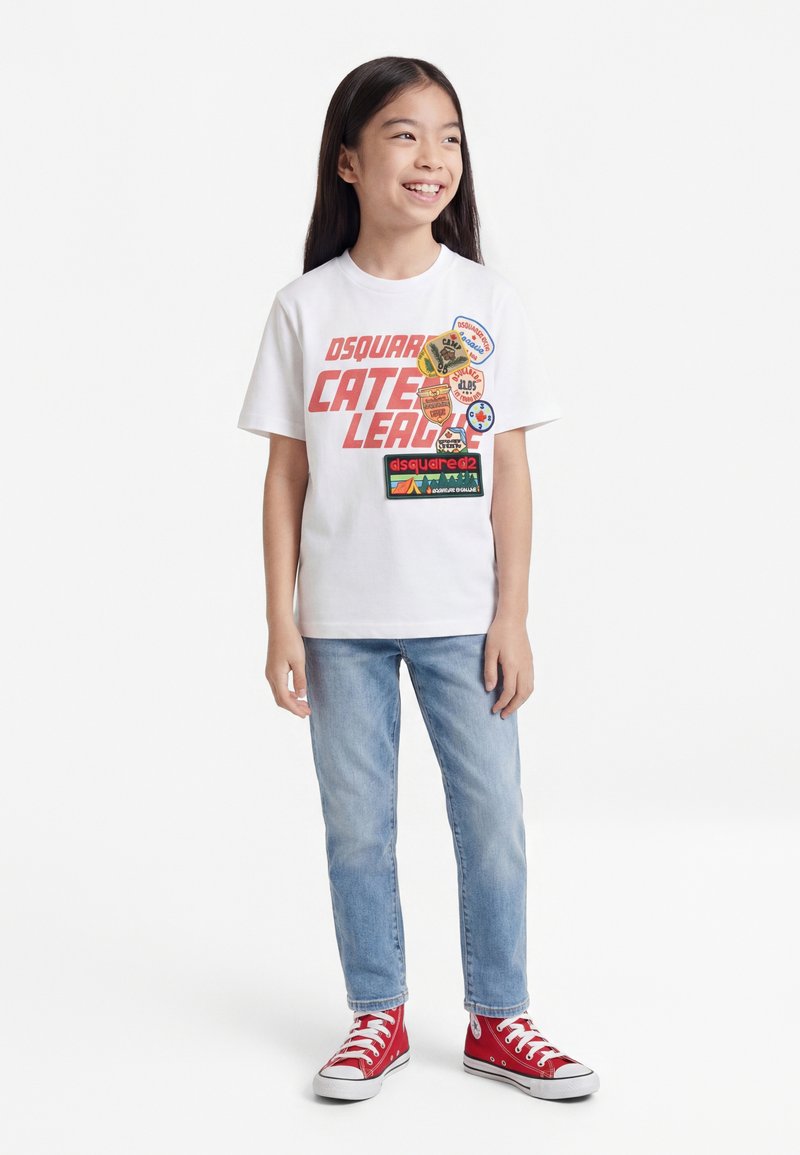 Jeune fille souriante, portant un t-shirt blanc à motifs avec des patches, un jean bleu clair et des baskets montantes rouges, debout sur fond blanc.