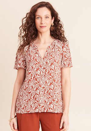 Donna con capelli lunghi e ricci che indossa una camicetta a maniche corte con bottoni, fantasia di foglie in ruggine e bianco, e pantaloni ruggine, in piedi davanti a uno sfondo semplice.