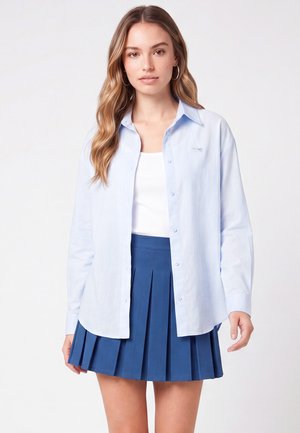 Giovane donna con capelli lunghi che indossa una camicia celeste con bottoni sopra una canottiera bianca e una gonna plissettata blu, in piedi su uno sfondo uniforme.