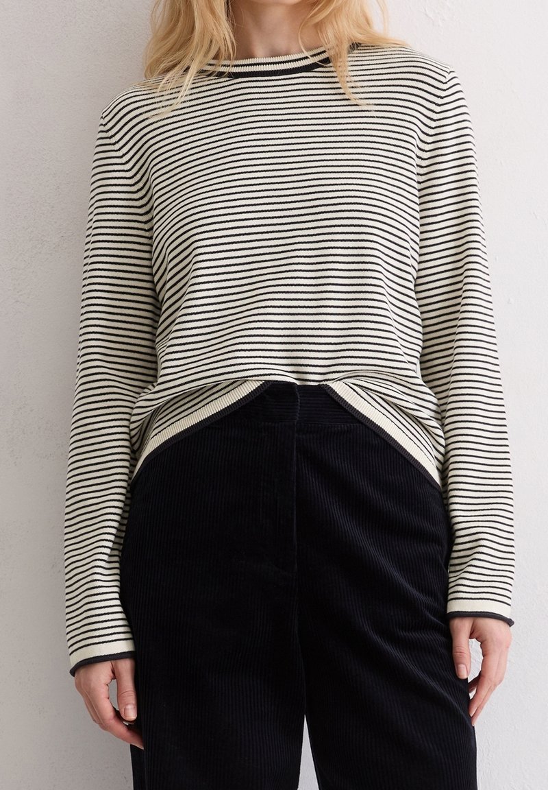 Femme portant un haut à manches longues rayé horizontalement blanc et noir, rentré dans un pantalon en velours noir taille haute.