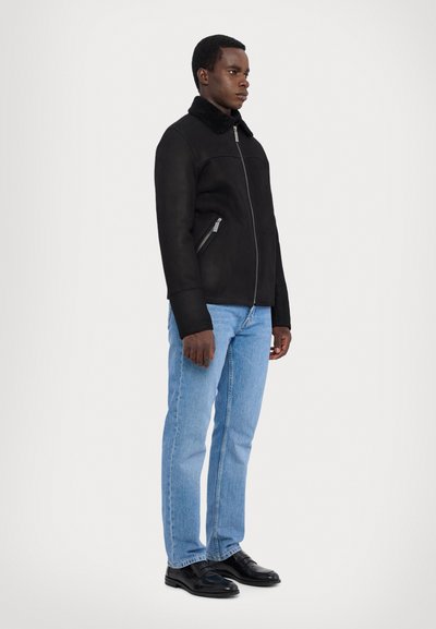Schwarze Wildlederjacke mit hohem Kragen, silbernem Reißverschluss und zwei schrägen Seitentaschen, kombiniert mit hellblauen geraden Jeans und schwarzen Schuhen.