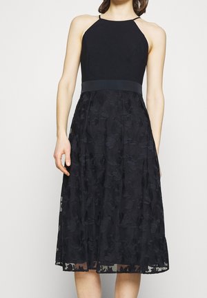 Halterneckklänning med en figurnära överdel, svart texturerat tyg och en utsvängd kjol med ett överlägg av blommönster.