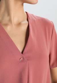 Blush pink bluse med V-hals og korte ærmer, præget af en enkelt lyserød knapdetalje foran og en struktureret stofoverflade.