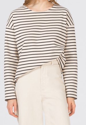 Nainen, joka pukeutunut pitkähihainen kerma-paita mustilla vaakaraitojen ja korkeavyötäröisiin kermahousuihin, yksiväristä taustaa vasten.