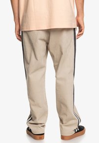 Beige cordfløyelsbukser med svarte sidelister, løs passform, elastisk midje og en glatt tekstur. Svarte sko med flat såle er synlige.