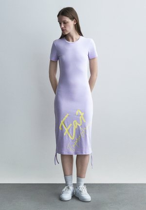 Mujer con un vestido midi ajustado color lavanda con mangas cortas y texto amarillo que dice "Eat Emperor Aenean", calzando zapatillas blancas y calcetines grises, con las manos detrás de la espalda.