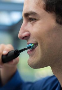 Spazzolino elettrico nero con testina blu e verde, tenuto da una persona mentre si lava i denti, che mostra un primo piano della bocca e della linea della mascella.