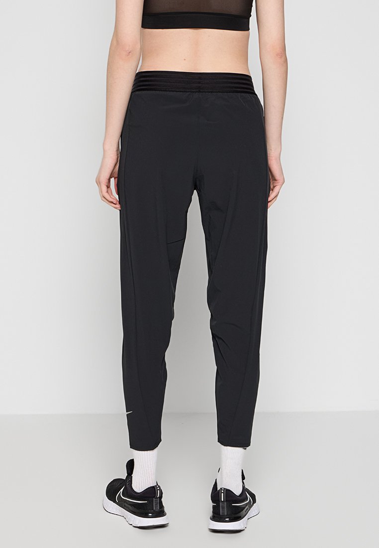 Personne portant un pantalon de sport noir, un crop top noir, des chaussettes blanches et des baskets Nike noires, se tenant dos à la caméra sur un fond uni.