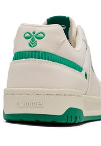 Beige Sneaker mit grünen Akzenten, strukturiertem Leder und einer glatten Sohle. Verfügt über ein grünes Logo und ein Streifendetail an der Ferse.
