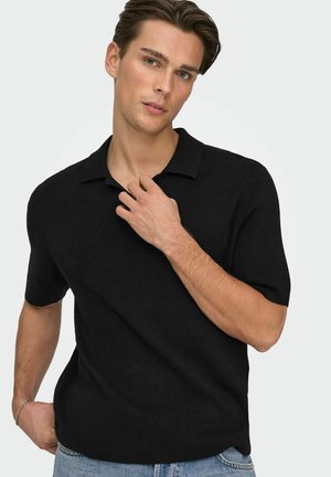 Mladý muž s krátkymi hnedými vlasmi v čiernej polokošeli s krátkym rukávom a modrých džínsoch, pózujúci s jednou rukou blízko goliera.