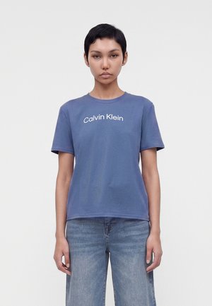 Personne aux cheveux courts noirs, portant un t-shirt bleu Calvin Klein et un jean bleu, debout devant un fond clair uni.
