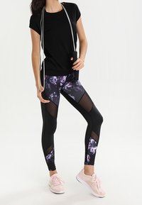 Zwart sport T-shirt gecombineerd met bloemenpatroon, zwarte en paarse leggings met mesh panelen. Lichtroze sneakers complementeren de outfit.