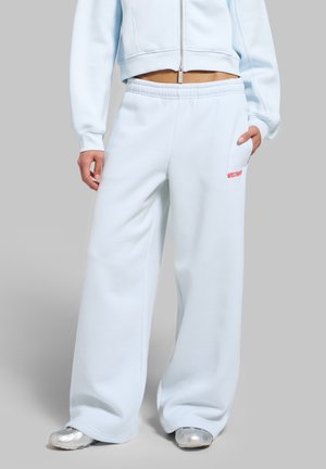 Personne portant un pantalon de survêtement bleu clair à jambes larges et une veste courte zippée assortie, les mains dans les poches, debout sur un fond gris.