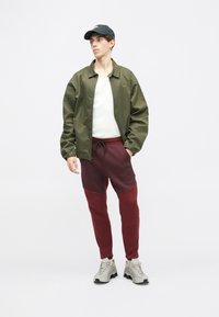 Olivzaļš viegls bomber jakas, balta garām piedurknēm T-kreklu, bordo sporta bikses ar tumšāku paneļu, pelēki čības ar teksturētu dizainu.