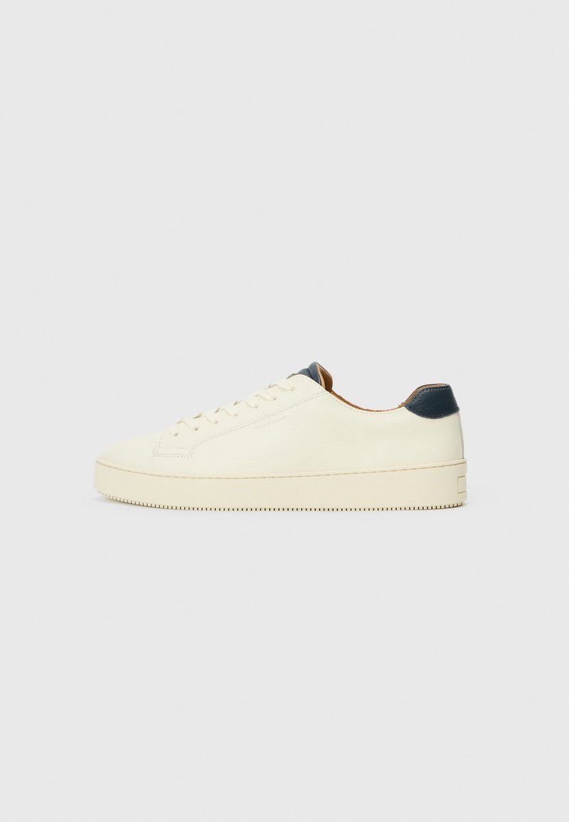 White leather sneaker with a textured surface, rounded toe, and a navy heel accent. Features side stitching and a textured sole.