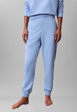 Persona che indossa pantaloni della tuta azzurri chiari e felpa abbinata, in piedi a piedi nudi su uno sfondo grigio uniforme.