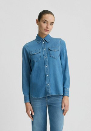 Camicia in denim blu con chiusura a bottoni, colletto e due tasche sul petto. Presenta dettagli di cucitura e maniche lunghe con polsini a bottoni.