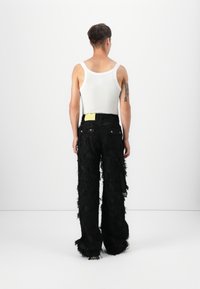 Schwarze, zerzauste Hose aus strukturiertem Stoff mit lockerer Passform, ausgestattet mit einem gelben Etikett, Gürtelschlaufen und hinteren Taschen. Getragen mit einem weißen Tanktop.