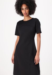 Rochie neagră cu mâneci scurte, confecționată dintr-un material fin, având un gât rotund și un corset ajustat care se lărgește ușor la tiv.