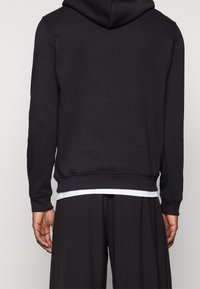 Sort hættetrøje med snøre i hætten, ribstrikkede manchetter og kant; lavet af blødt stof. Bruges over en hvid undertrøje. Rent design, ingen synlige logoer.