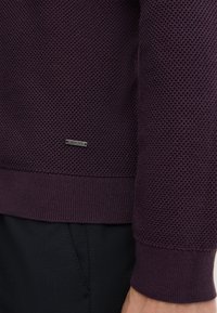 Gestrickter Pullover in Dunkelviolett, mit einem strukturierten Muster und gerippten Bündchen. Silbernes Logo-Tag in der Nähe des unteren Saums platziert.
