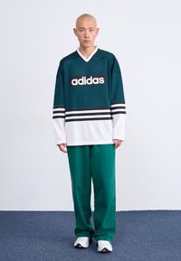 Muškarac stoji u zelenoj i bijeloj Adidas majici, usklađenim zelenim hlačama i bijelim tenisicama na jednostavnoj svijetloj pozadini.