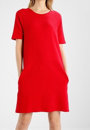 Frau trägt ein locker sitzendes, kurzärmeliges rotes Kleid mit den Händen in den Taschen, steht vor einem schlichten, hellen Hintergrund.