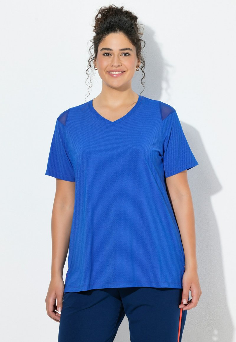 Žena s kudrnatými vlasy svázanými nahoru, oblečená v jasně modrém krátkém topu s výstřihem do V a tmavě modrých kalhotách s červeným pruhem, usmívající se do kamery.