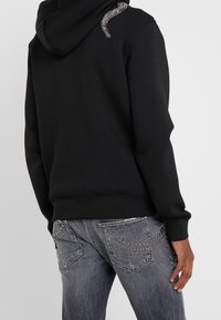 Sudadera negra con un detalle de serpiente texturizada en el hombro derecho, combinada con jeans grises con bolsillos traseros con tachuelas metálicas.