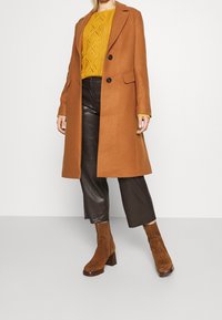 Brun ullrock med snedkrage och två knappar, tillsammans med en gul stickad tröja och bruna läderbyxor. Bruna mocka ankelstövlar.