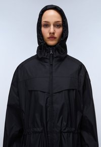 Giacca nera impermeabile con cappuccio, dotata di chiusura con zip, cordino intorno al viso e elastiche in vita per un look aderente.