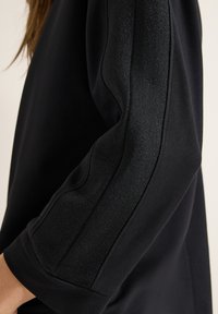 Mustapaita, jossa on sileä kangas ja teksturoitu, kiiltävä yksityiskohta hihojen varrella. Muotoilu on yksinkertainen ja rento leikkaus.