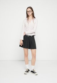 Ανοιχτό ροζ ριγέ μπλούζα με μακριά μανίκια και ζώνη στη μέση, συνδυασμένη με μαύρη πλισέ φούστα, λευκές κάλτσες και μαύρα αθλητικά παπούτσια.