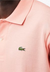 Polo shirt rose en tissu doux, avec un logo crocodile brodé vert sur la poitrine gauche et une patte de boutonnage à trois boutons.