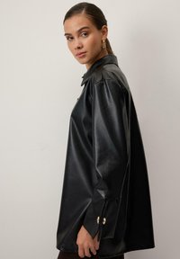Sort faux læder skjorte med krave, løs pasform og knaplukning ved håndleddene. Glat tekstur med en let glans og skræddersyede designdetaljer.