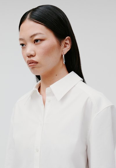 White button-up shirt with a collar, featuring a smooth texture and structured fit, complemented by a large silver hoop earring.