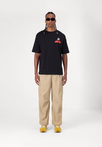 Μαύρη oversized μπλούζα T-shirt με κόκκινο γραφικό "NEW ERA", μπεζ cargo παντελόνι και κίτρινα sneakers με λευκές λεπτομέρειες. Μοντέλο που φοράει γυαλιά ηλίου.