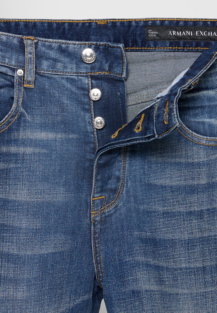 Jeans in denim blu con chiusura a bottoni, bottoni argento a vista, cuciture gialle e etichetta Armani Exchange all'interno della cintura.