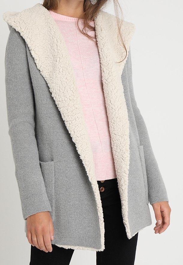 Femme portant un manteau gris ouvert devant avec une doublure en polaire blanche sur un pull rose clair et un pantalon noir.