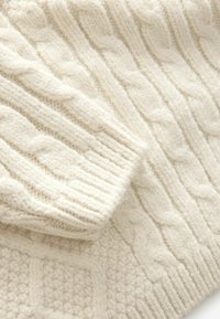 Tissu tricoté couleur crème avec motifs torsadés et côtelés, présentant les détails des manches et du corps d'un pull chaud texturé.