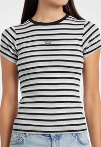 Žena nosi usku crno-bijelu horizontalno prugastu majicu s logom "GUESS JEANS" na prsima i svijetloplave traperice.